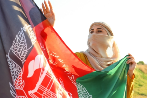 [ai] A woman wearing a light-colored scarf holds the Afghan flag, which features the national emblem, in a field during sunset. She is waving the flag, showcasing a sense of pride in her heritage.